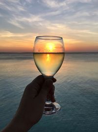 Man holding glass against sea during sunset
