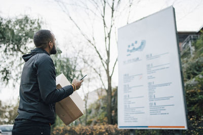 Male worker with box and smart phone looking at billboard during delivery