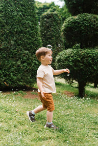 Cute happy little baby boy is running on the grass in the park trying to catch a soap bubble