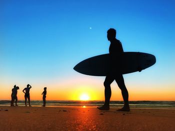 Full length of man with surfboard at beach against blue sky during sunset