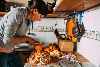 Side view of adult housewife chopping fresh pumpkin on table with various vegetables while preparing healthy natural cream soup at home
