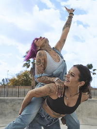 Low angle of delighted woman with pink hair piggybacking cheerful female friend with tattoos while having fun together at weekend
