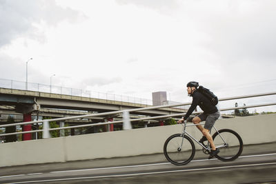 Side view of male commuter with backpack riding bicycle on bridge against sky
