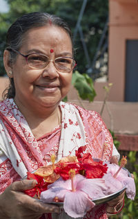 An aged bengali woman offering prayer to sun god, holding bouquet of hibiscus flowers in tray.