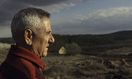 Side view of thoughtful senior man standing on field against cloudy sky