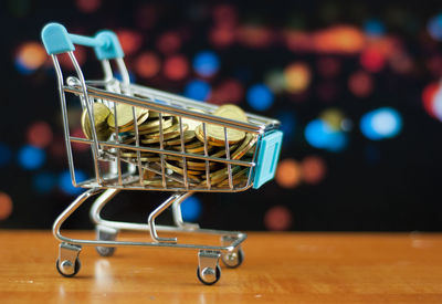 Close-up of miniature shopping cart with coins on table against lights