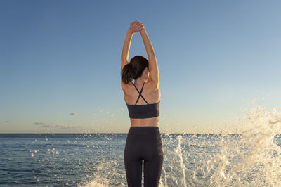 Fit female doing an arm stretch warm up exercise by the sea with waves splashing.