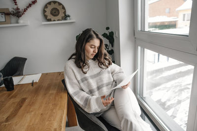 Woman reading document while sitting on armchair near window at home