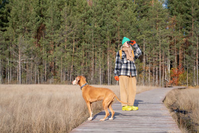 Woman with hunting dog walking in ecological trail in autumn forest looking through binoculars.