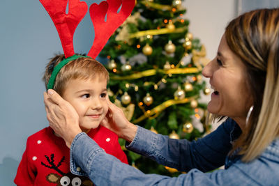 Smiling mother helping son in wearing headband against christmas tree
