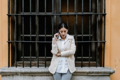 Confident adult hispanic female with dark hair and crossed arms in smart casual outfit standing near aged building with metal window bars and talking smartphone