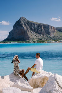 People sitting on rock by sea against mountains