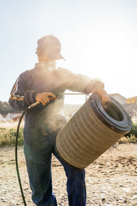 Unrecognizable farmer in workwear and protective respirator mask spraying water from hose to dirty filter of agricultural machine during maintenance work in farm yard