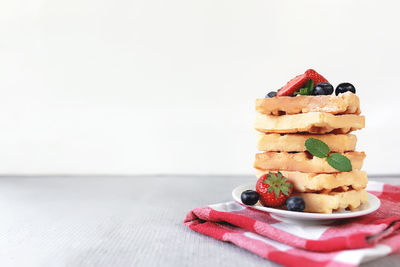 Stack of waffles on a white plate on the towel and table with blueberry, chopped strawberry and mint