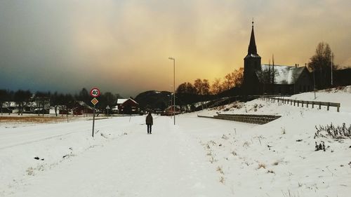 Rear view of man standing on snow covered field by church against sky during sunset