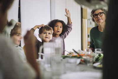 Children raising hands while sitting with female teacher during lunch time at kindergarten