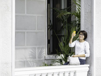 Woman makes selfie on smartphone. pretty female sits on balcony with palm trees in flower pots.