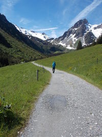 Rear view of man walking on road amidst field towards mountain range