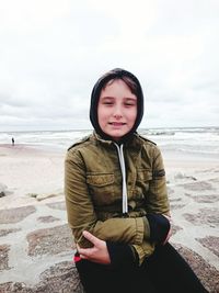 Portrait of girl in warm clothing sitting on rock at beach against cloudy sky