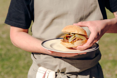 Unrecognizable male cook in apron standing in countryside with plate of appetizing burger