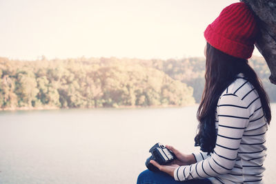 Side view of woman with camera by lake against clear sky