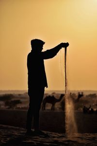 Silhouette man pouring sand while standing at desert against sky during sunset