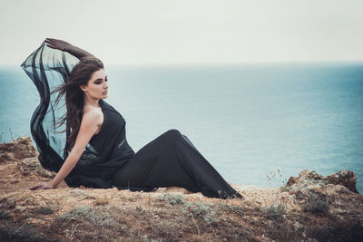 Young woman sitting on land by sea against sky