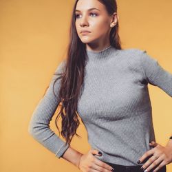 Thoughtful young woman with hands on hip looking away against yellow background