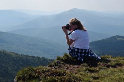Rear view of woman photographing while sitting on mountain against sky