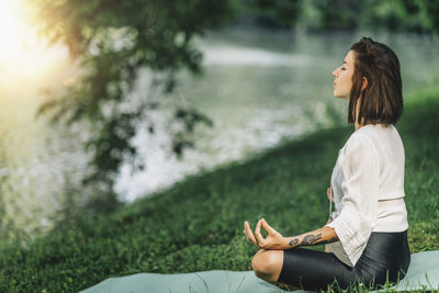 Young woman sitting in lotus position and practicing meditation near water in the nature