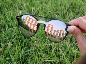 Cropped hand holding sunglasses with reflection of old ruins over grassy land