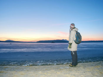 Woman in warm long jacket stay on sandy beach at frozen lake or river. winter chilly morning 