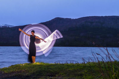 Woman spinning illuminated lightning equipment while standing by lake against sky at night