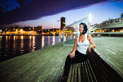 Woman sitting on bench against sky in city at dusk