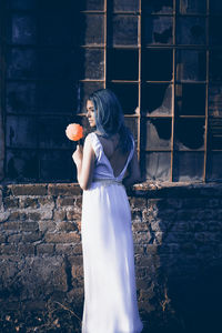 Woman holding flower while standing by brick wall