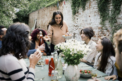 Smiling woman holding bowl while talking to male and female friends during dinner party in back yard