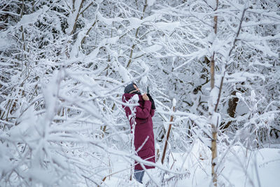 A girl in a burgundy down jacket photographs a snow-covered forest on a mobile phone. person