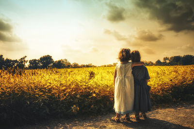 Rear view of siblings standing on field against sky during sunset