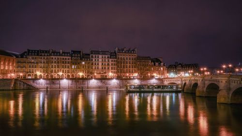 Illuminated bridge over river by buildings against sky at night