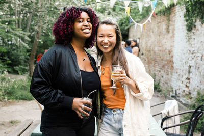 Portrait of happy multiracial female friends with drinks standing together during party in back yard