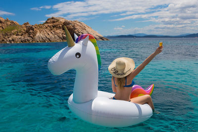 Rear view of woman sitting on inflatable unicorn in aegean sea against sky