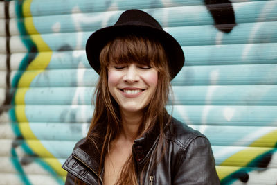 Smiling woman with eyes closed wearing hat against graffiti shutter in city