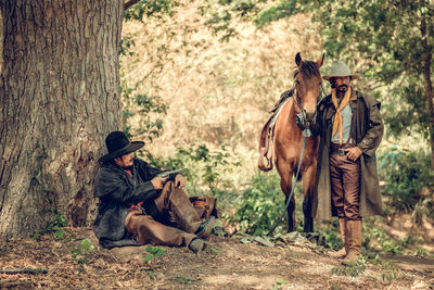 Man with horse standing by friend sitting in forest