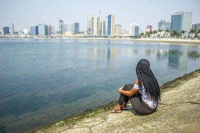 Full length of woman sitting on riverbank in city against sky