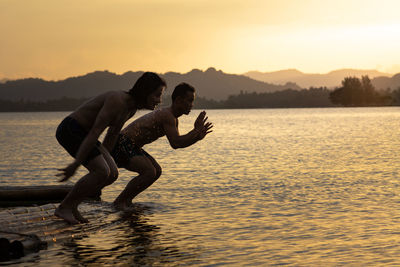 Shirtless men jumping in lake against sky during sunset