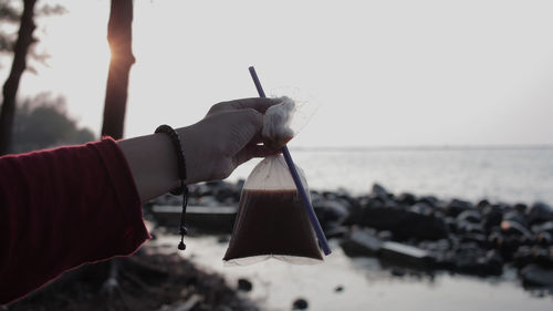 Cropped hand holding drink in plastic bag with straw by sea