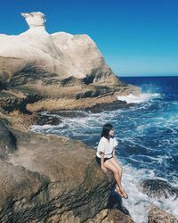 Young woman sitting on rock formation while looking at sea against sky