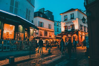 People walking on street amidst buildings in city at dusk