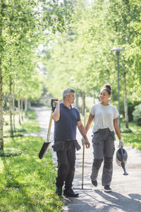 Full length of female trainee with male instructor holding gardening equipment and walking footpath at garden