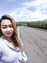 Portrait of smiling young woman standing on road against sky